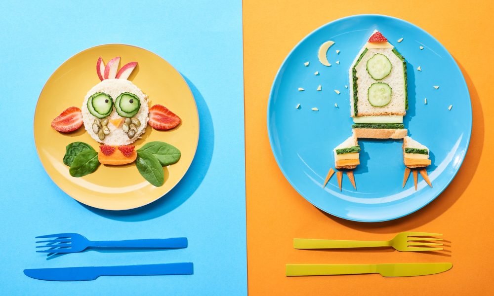 top view of plates with fancy face and rocket made of food for childrens breakfast near cutlery on